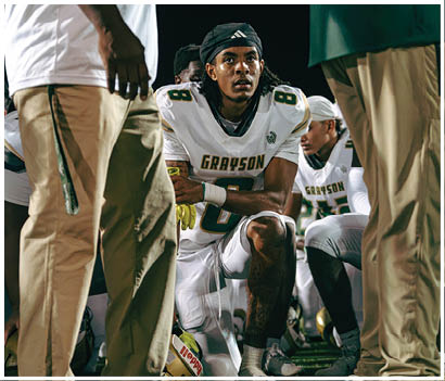 Two people in white and green football uniforms stand on a field with one clenching a fist and a referee and opposing players in red uniforms visible in the background. Description generated by AI