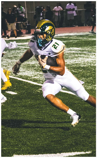 A person in a white and green football uniform is running with the ball on a field while two people in red uniforms approach from either side. Description generated by AI