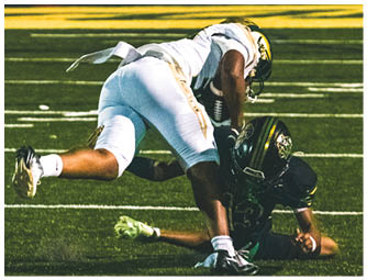 A person wearing green sports shorts with the word “GRAYSON" on the side and white gloves stands on a football field with other players in the background. Description generated by AI