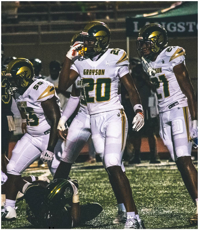 Two people in football uniforms with helmets stand on a field, one facing forward and the other facing away, with a third person in casual clothing in the background. Description generated by AI