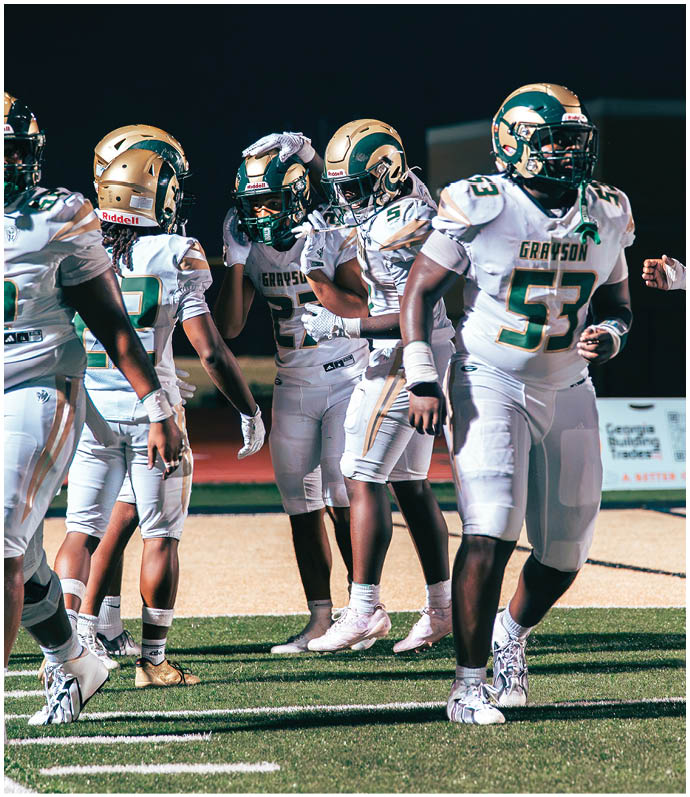 Two people in football uniforms and helmets are on a field with a referee standing behind them. Description generated by AI