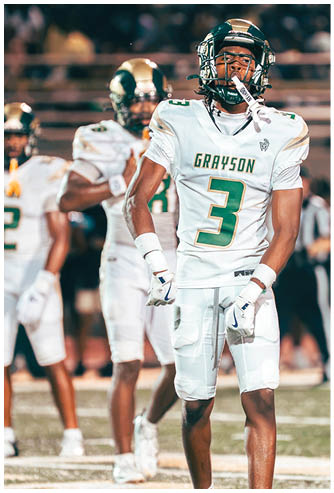 A person in a white and green football uniform with the name Grayson is running on a field holding a football while another person in similar attire stands nearby and a photographer captures the moment in the background. Description generated by AI