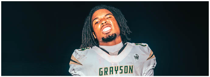 A person wearing a white football uniform with “GRAYSON" on the chest is running while holding a football on a field. Description generated by AI