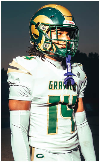 Two people in football uniforms with helmets stand on a field during a game, with one person facing forward and the other partially visible from behind. Description generated by AI