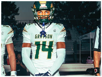 A person wearing a white and green football uniform with the word Grayson on the pants is running on a football field during a game with other players and a crowd in the background. Description generated by AI