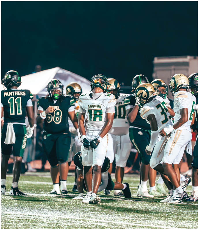 Several people in football uniforms are lined up on a field facing each other, preparing for a play during a game with spectators in the background. Description generated by AI