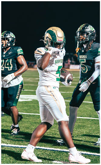Two football players in green and white uniforms are running on a field with a referee standing behind them. Description generated by AI
