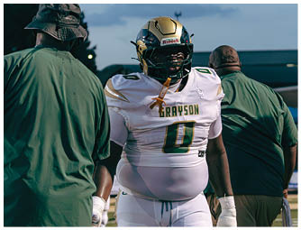 A person wearing green football pants and a white jersey with the number 11 stands on a football field with another person in red football gear in the background. Description generated by AI