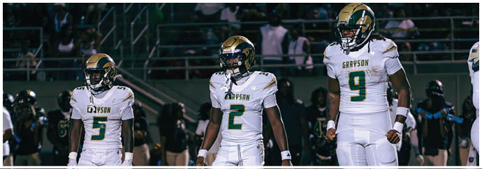 Two people wearing football helmets and white jerseys are standing on a field with a blurred crowd in the background. Description generated by AI