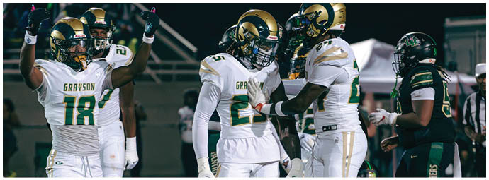 Three people in football uniforms are positioned on a field, with one person in a white jersey and green pants bent forward in a ready stance between two others wearing green and red pants. Description generated by AI