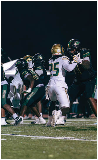 Two people in football uniforms and helmets are positioned on a field with one crouching and holding a football while the other stands behind. Description generated by AI