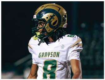 A person wearing green sports shorts with the word “GRAYSON" on the side and white gloves stands on a football field with other players in the background. Description generated by AI