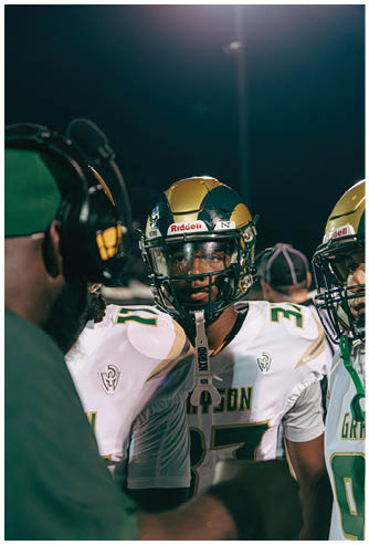 A person wearing a football helmet is in the foreground with a stadium and a scoreboard reading “Sandy Creek State Champions" in the background during sunset. Description generated by AI