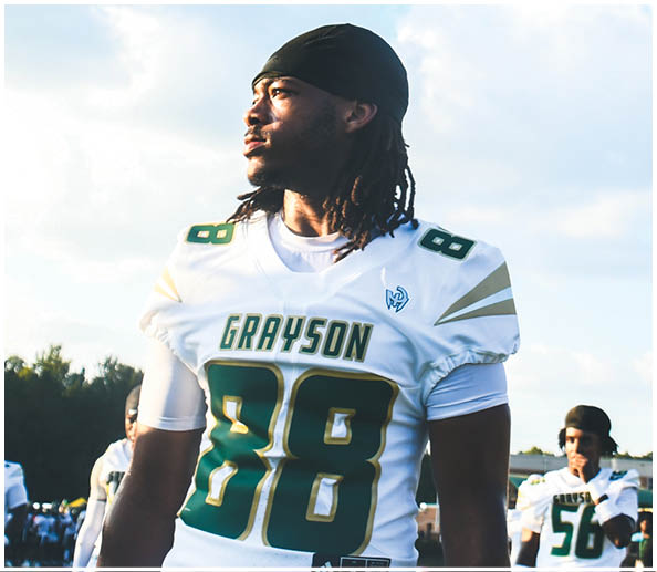 A person in a white and gold football uniform is crouched in an athletic stance on a green football field with other players and a coach in the background. Description generated by AI