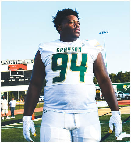 A person with long dreadlocks wearing a white football jersey with the name Grayson and the number 28 stands in front of two other people in similar football uniforms and helmets. Description generated by AI