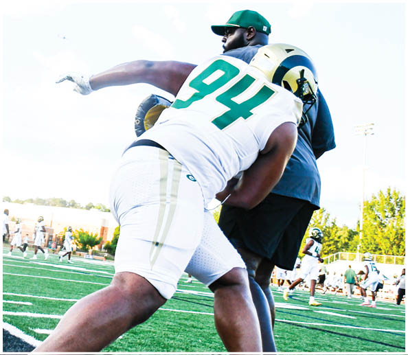 A person in a white and green football uniform with a gold helmet is on a field facing away from the camera while another person in a maroon and gold uniform stands in the background near empty bleachers. Description generated by AI