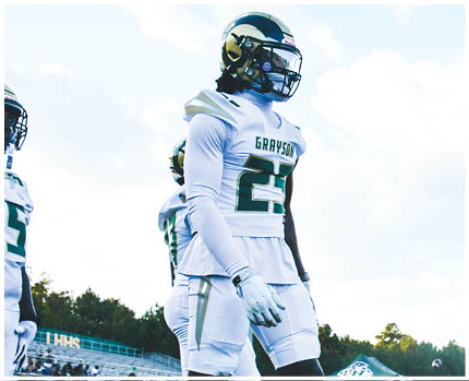 A person wearing a white and gold football uniform with the number 20 stands on a field while two other people in red and gold football uniforms are visible in the background near empty bleachers. Description generated by AI