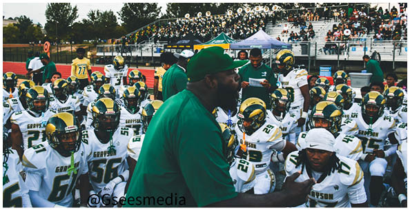 A person wearing a white sports uniform with the number 47 is standing on a field with other people in similar uniforms and spectators seated in bleachers in the background. Description generated by AI