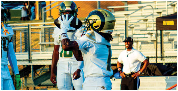 Four people wearing white football uniforms with gold helmets and large numbers on their backs are standing on a field. Description generated by AI