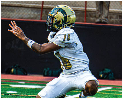 A person wearing a white football uniform with a gold and black helmet is kneeling on a green football field with arms extended forward. Description generated by AI