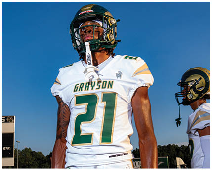A person in a white and gold football uniform is crouched on a field with other players in similar uniforms standing in the background. Description generated by AI