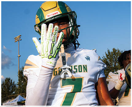 Two people wearing football helmets and white jerseys with gold and green accents are facing each other in a stadium with empty bleachers in the background. Description generated by AI