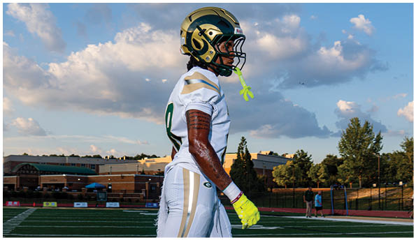 Four people wearing football helmets and uniforms are lined up facing an opponent on a field. Description generated by AI