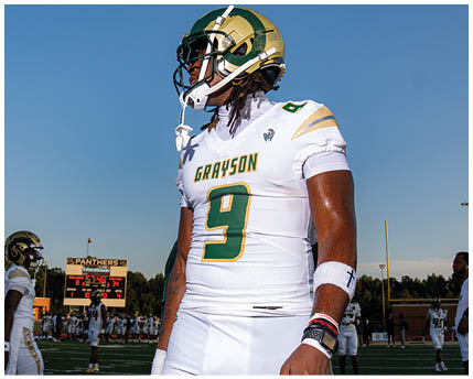 A person wearing sunglasses and a headset is speaking intensely while another person in a football helmet looks on in the background. Description generated by AI