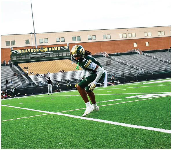 A person in a white and gold football uniform is crouched in an athletic stance on a green football field with other players and a coach in the background. Description generated by AI