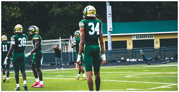 Two people wearing white sports uniforms with green and yellow stripes stand on a field with others in the background. Description generated by AI