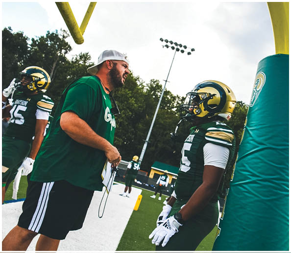 A person in a white and green football uniform with a gold helmet is on a field facing away from the camera while another person in a maroon and gold uniform stands in the background near empty bleachers. Description generated by AI