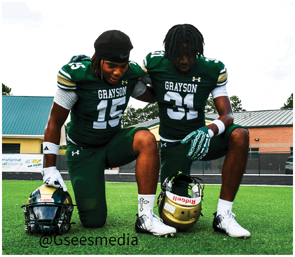 A person in a white and gold football uniform is sitting on the field holding a football while other players stand in the background. Description generated by AI