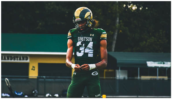 A person wearing a white football uniform and helmet is running with a football on a field while people in green cheerleading outfits stand in the background. Description generated by AI