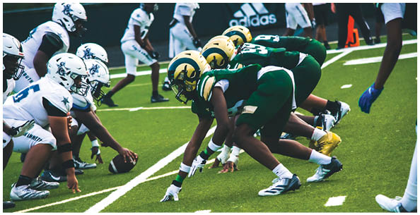 Four people wearing white football uniforms with gold helmets and large numbers on their backs are standing on a field. Description generated by AI
