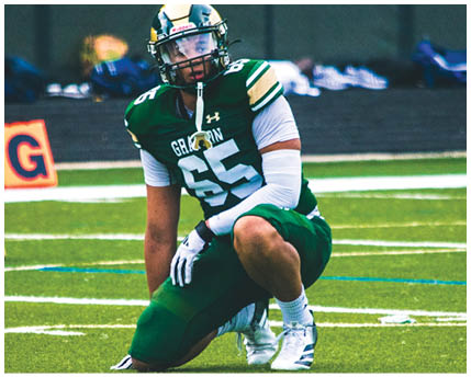 A person wearing a white football uniform with a gold and black helmet is kneeling on a green football field with arms extended forward. Description generated by AI