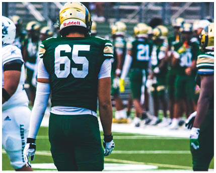 A person in a white and gold football uniform is crouched on a field with other players in similar uniforms standing in the background. Description generated by AI