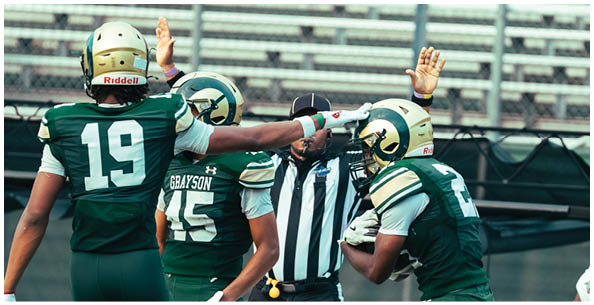 Four people wearing football helmets and uniforms are engaged in conversation on a field with bleachers in the background. Description generated by AI