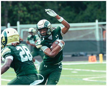 Two people wearing football helmets and white jerseys with gold and green accents are facing each other in a stadium with empty bleachers in the background. Description generated by AI