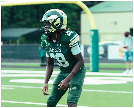 A person wearing sunglasses and a headset is speaking intensely while another person in a football helmet looks on in the background. Description generated by AI