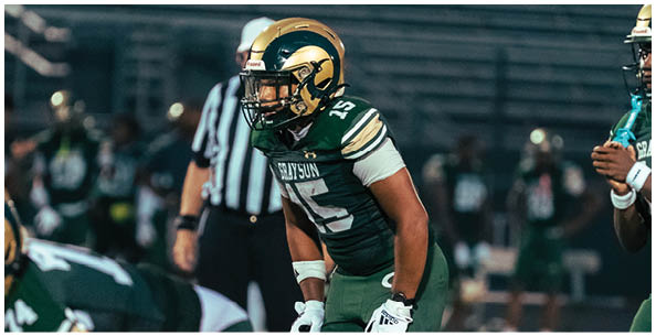Two people wearing football helmets and uniforms face each other on a football field with cheerleaders and spectators in the background. Description generated by AI