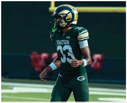 Two people in white and gold football uniforms tackle a person in a maroon and gold football uniform holding a football near a goalpost on a field. Description generated by AI