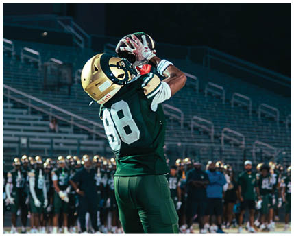 A person in a white football uniform with “GRAYSON" on the jersey is celebrating on a football field while two other players in maroon and gold uniforms are in the background. Description generated by AI