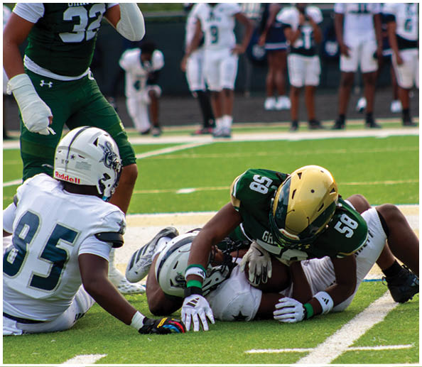 A person wearing a green sweatshirt and white cap is holding papers and appears to be speaking on a sports field while a player in a white and gold football uniform stands nearby. Description generated by AI