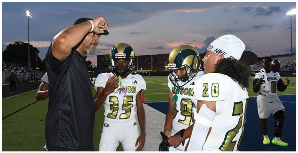 People wearing green and white football uniforms stand on a field while a person in an orange shirt and dark pants holds a whistle and appears to be coaching. Description generated by AI