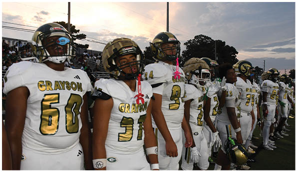 Two people in green and white football uniforms with helmets are on a football field, with one person appearing to hold or block the other while a person in a green shirt watches nearby. Description generated by AI