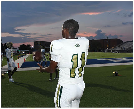 A person wearing a green and white football uniform is running on a field while holding a football, with another person closely behind. Description generated by AI