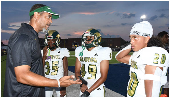 Several people in green and white football uniforms are positioned on a grassy field, preparing for a play. Description generated by AI