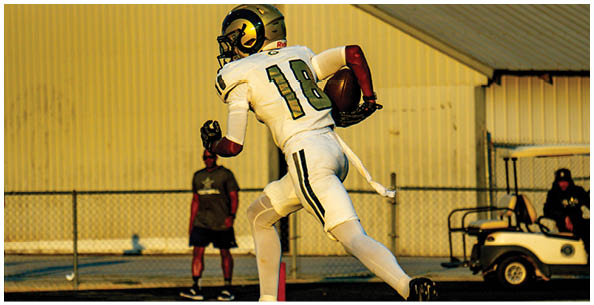 A person wearing a green and white football uniform is crouched on a field with empty bleachers and trees in the background. Description generated by AI