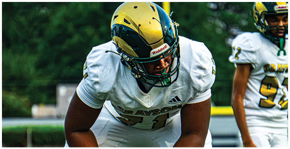Four people in football uniforms and helmets are lined up in a ready stance on a field, preparing for a play. Description generated by AI