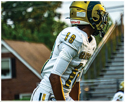 Two people in football uniforms are jumping to catch a ball on a field while another person stands nearby holding a drink and smiling. Description generated by AI